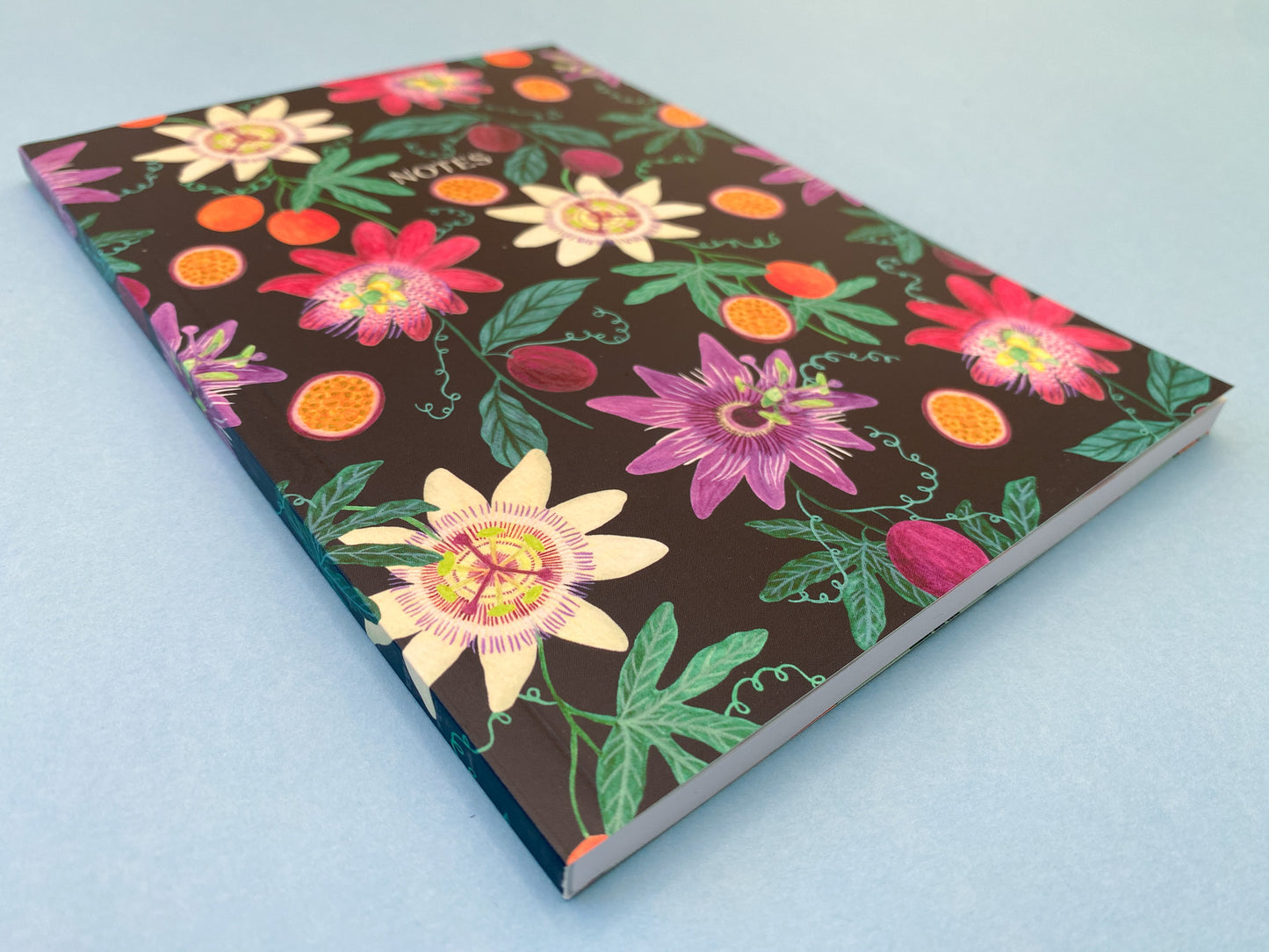 Close up details. Passion fruit and flowers A5 notebook. Dark blue background with pale yellow, dark pink, and purple passion flowers. With orange and purple passion fruits and green foliage.