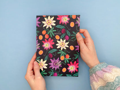 Passion fruit and flowers A5 notebook. Dark blue background with pale yellow, dark pink, and purple passion flowers. With orange and purple passion fruits and green foliage.