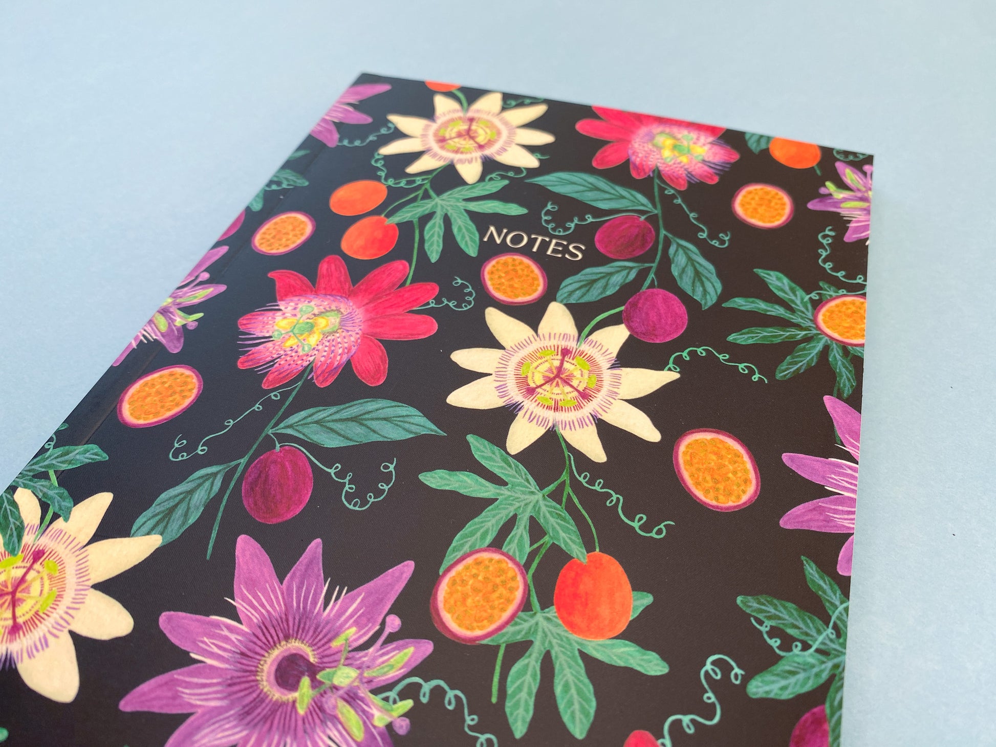 Close up details. Passion fruit and flowers A5 notebook. Dark blue background with pale yellow, dark pink, and purple passion flowers. With orange and purple passion fruits and green foliage.