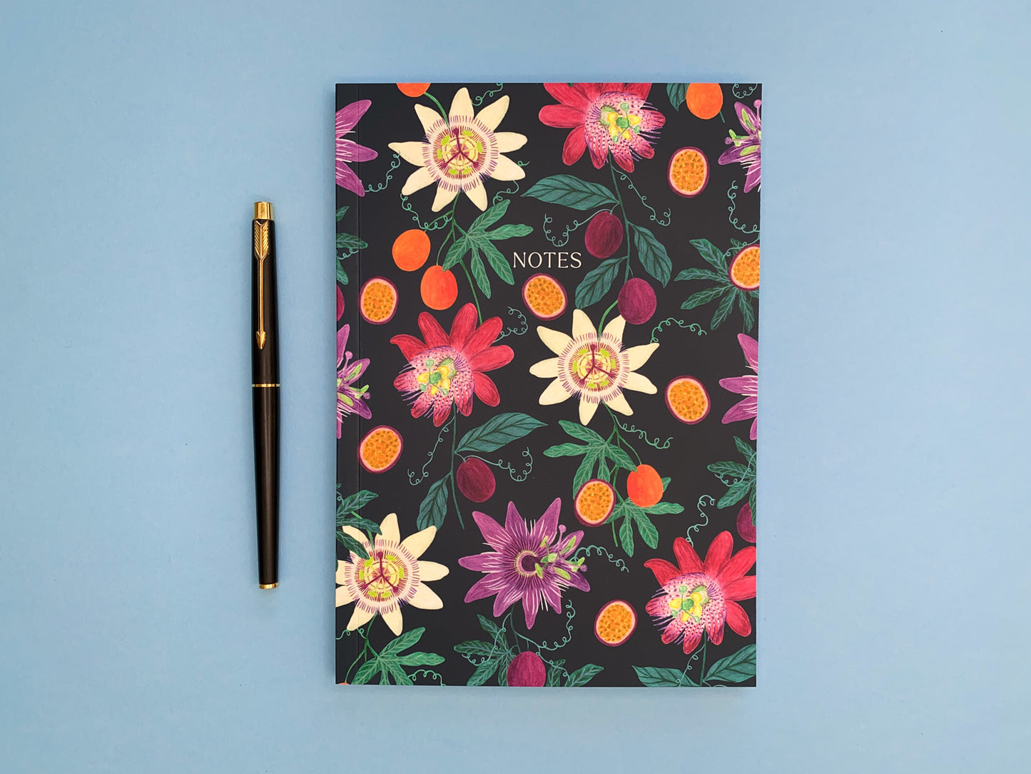 Passion fruit and flowers A5 notebook. Dark blue background with pale yellow, dark pink, and purple passion flowers. With orange and purple passion fruits and green foliage.