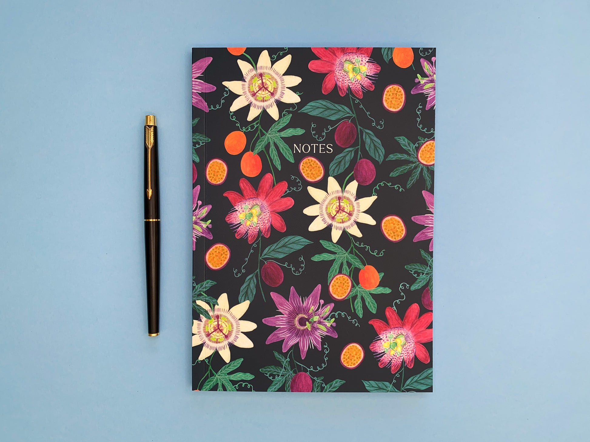 Passion fruit and flowers A5 notebook. Dark blue background with pale yellow, dark pink, and purple passion flowers. With orange and purple passion fruits and green foliage.