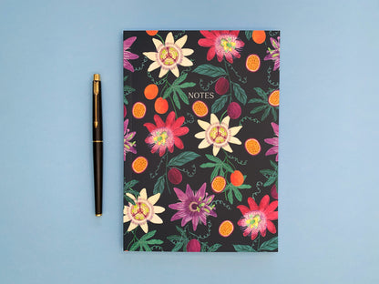Passion fruit and flowers A5 notebook. Dark blue background with pale yellow, dark pink, and purple passion flowers. With orange and purple passion fruits and green foliage.