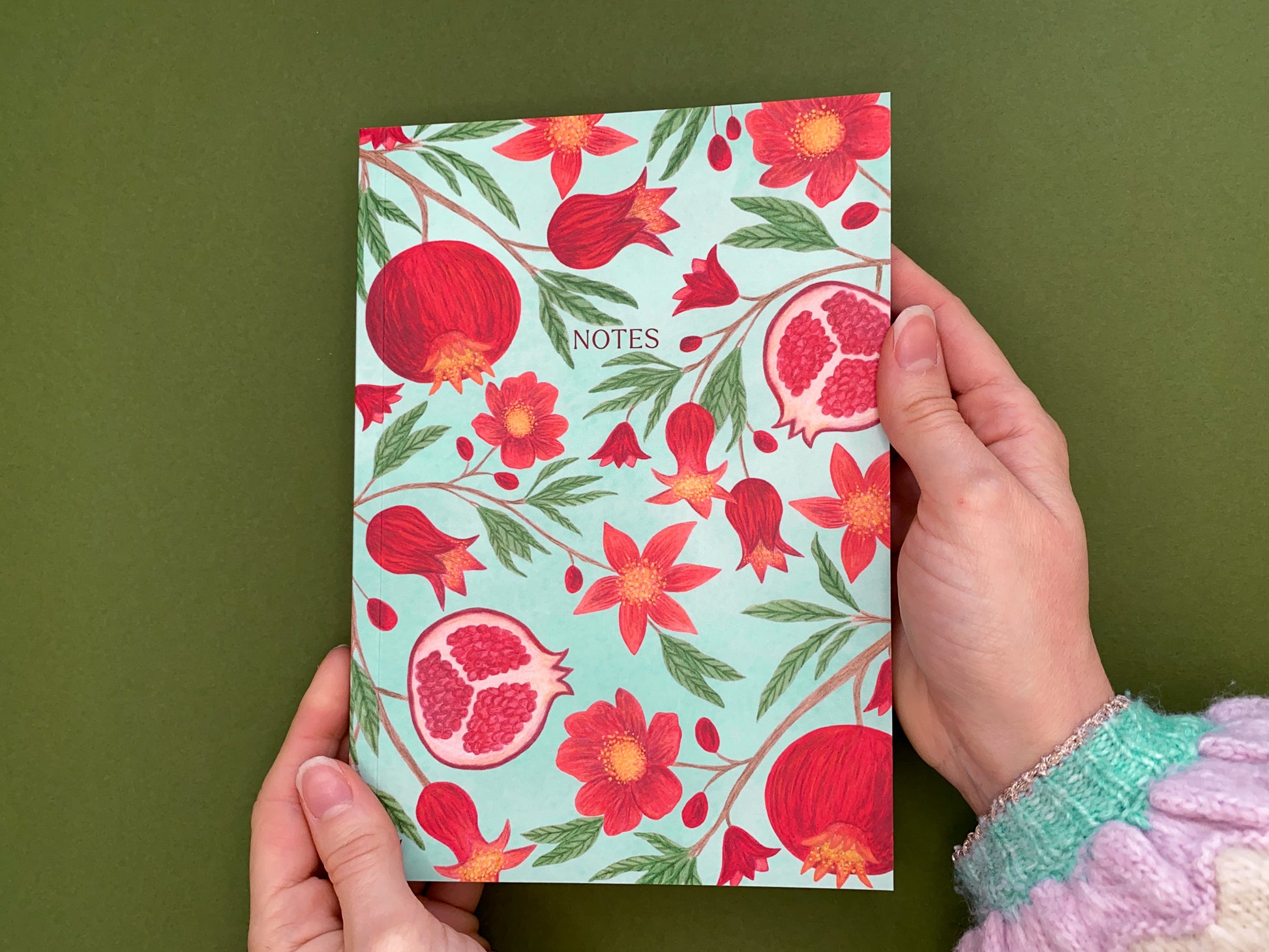 Pomegranate A5 notebook. Pale green watercolour background. Vibrant red pomegranates and flowers pattern with green leaves. The text notes added on the front cover. 