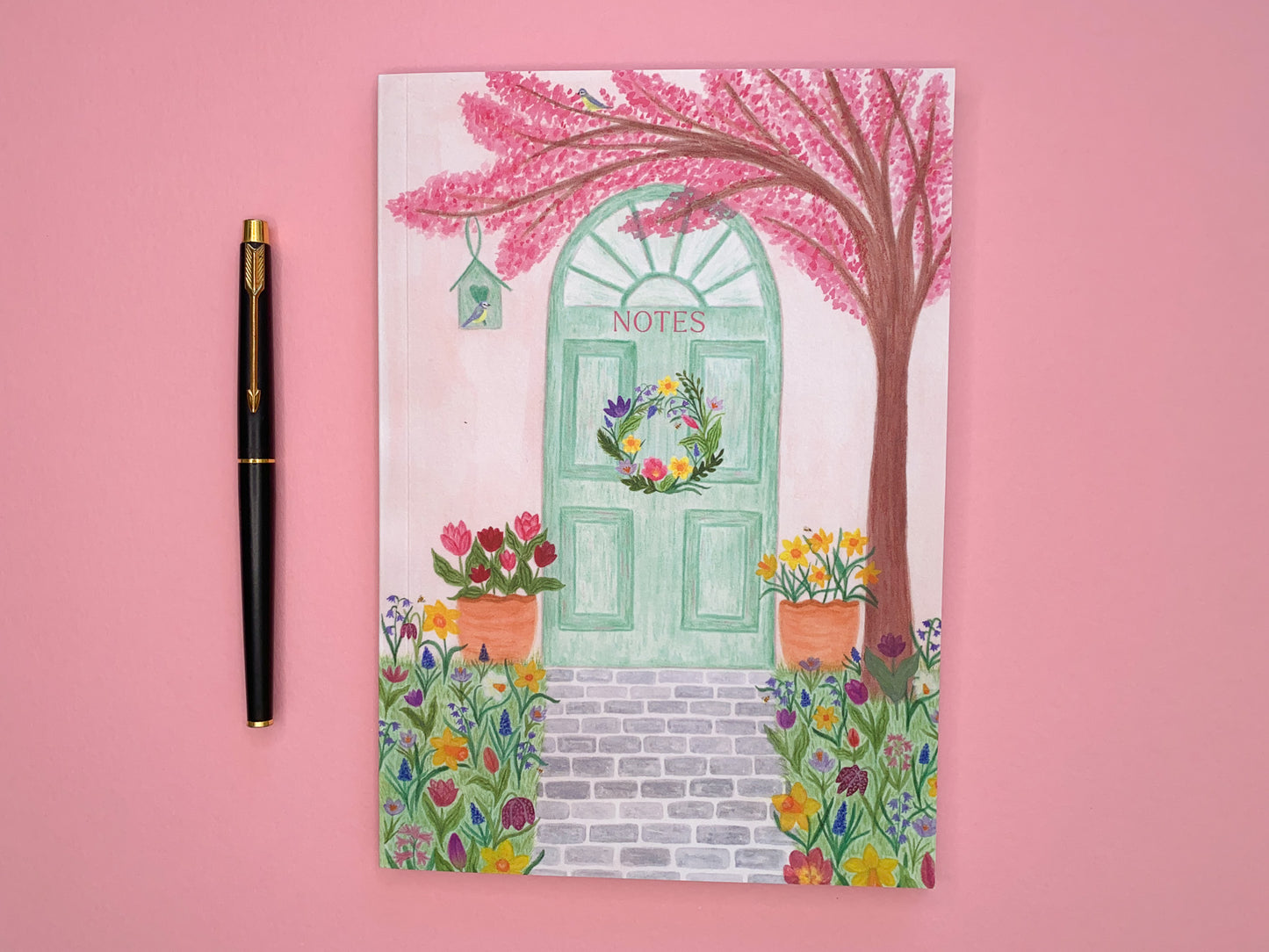 Spring door A5 notebook. Softcover perfect bound and lined inside pages. Front cover illustration of spring pastel green door with a wreath. Pale pink wall. Pink cherry blossom tree with blue tits and bird house. Tulips and daffodils in pots on either side of the door. Grey brick pavement in the middle. Spring meadow flowers on both side of pavement. Flowers include daffodils, tulips, bluebells, hyacinths, Fritillary,  crocuses and bees. Notes text placed on green door.