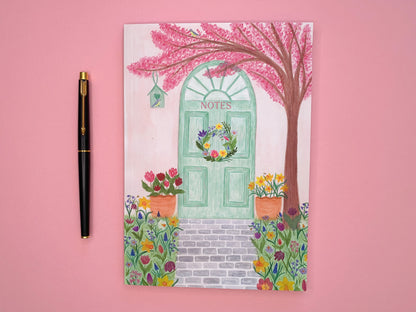 Spring door A5 notebook. Softcover perfect bound and lined inside pages. Front cover illustration of spring pastel green door with a wreath. Pale pink wall. Pink cherry blossom tree with blue tits and bird house. Tulips and daffodils in pots on either side of the door. Grey brick pavement in the middle. Spring meadow flowers on both side of pavement. Flowers include daffodils, tulips, bluebells, hyacinths, Fritillary,  crocuses and bees. Notes text placed on green door.