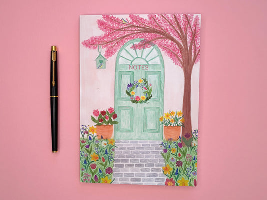 Spring door A5 notebook. Softcover perfect bound and lined inside pages. Front cover illustration of spring pastel green door with a wreath. Pale pink wall. Pink cherry blossom tree with blue tits and bird house. Tulips and daffodils in pots on either side of the door. Grey brick pavement in the middle. Spring meadow flowers on both side of pavement. Flowers include daffodils, tulips, bluebells, hyacinths, Fritillary,  crocuses and bees. Notes text placed on green door.