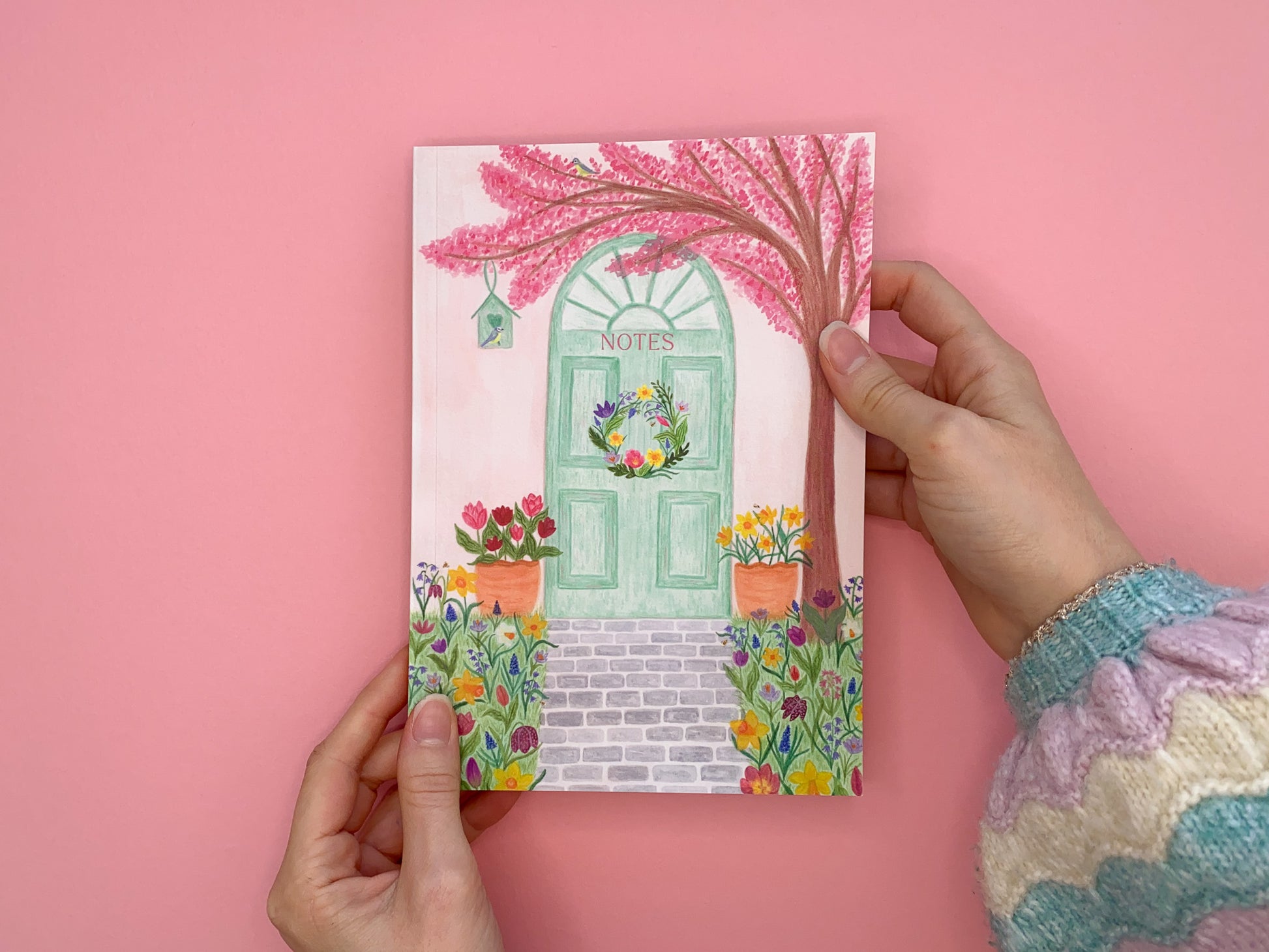 Spring door A5 notebook. Softcover perfect bound and lined inside pages. Front cover illustration of spring pastel green door with a wreath. Pale pink wall. Pink cherry blossom tree with blue tits and bird house. Tulips and daffodils in pots on either side of the door. Grey brick pavement in the middle. Spring meadow flowers on both side of pavement. Flowers include daffodils, tulips, bluebells, hyacinths, Fritillary,  crocuses and bees. Notes text placed on green door.