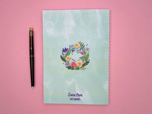 Back of spring door notebook. Pale green watercolour background with the spring wreath in the middle. Wreath includes daffodils, tulips, bluebells, crocuses, green foliage, bees and a blue tit bird.  
