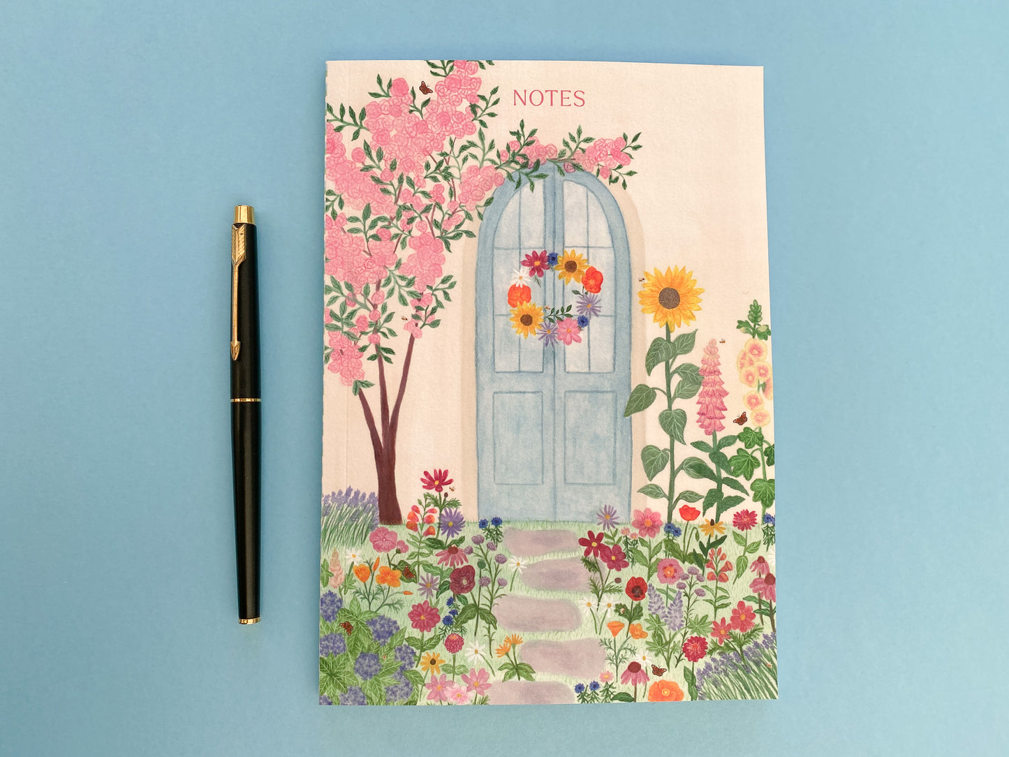 Summer blue door A5 notebook. Pale yellow wall. Pastel blue door with summer wreath. Pink rose bush tree on left. Sunflower, foxglove and hollyhock on the right. Grey stepping stones towards the door with summer flowers on either side and grass. Flowers include, lavender, poppies, cosmos, cornflowers, dahlias, echinacea, daises, lupins, hydrangea, zinnias, snapdragons, coneflowers, asters, verbena with bees and peacock butterflies. The text notes is placed at the top of notebook above the door.