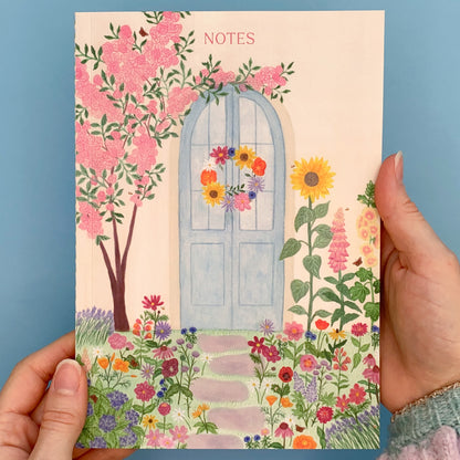 Summer blue door A5 notebook. Pale yellow wall. Pastel blue door with summer wreath. Pink rose bush tree on left. Sunflower, foxglove and hollyhock on the right. Grey stepping stones towards the door with summer flowers on either side and grass. Flowers include, lavender, poppies, cosmos, cornflowers, dahlias, echinacea, daises, lupins, hydrangea, zinnias, snapdragons, coneflowers, asters, verbena with bees and peacock butterflies. The text notes is placed at the top of notebook above the door.