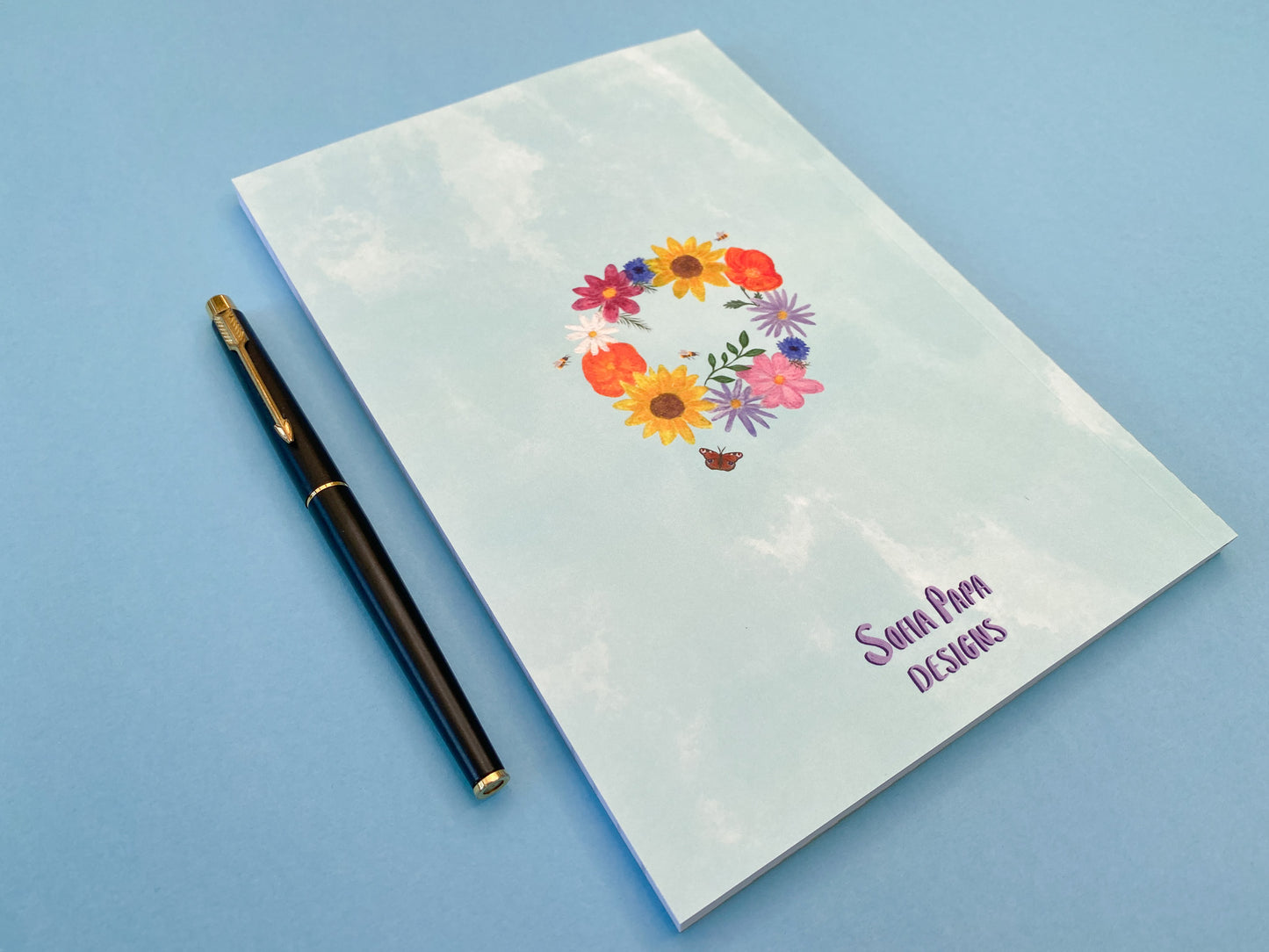 The back cover design. Pale blue watercolour background with summer wreath in the middle. Flowers include pink cosmos, blue cornflowers, Sunflowers,  orange poppies, purple asters, daisy, green foliage, bees and a peacock butterfly.