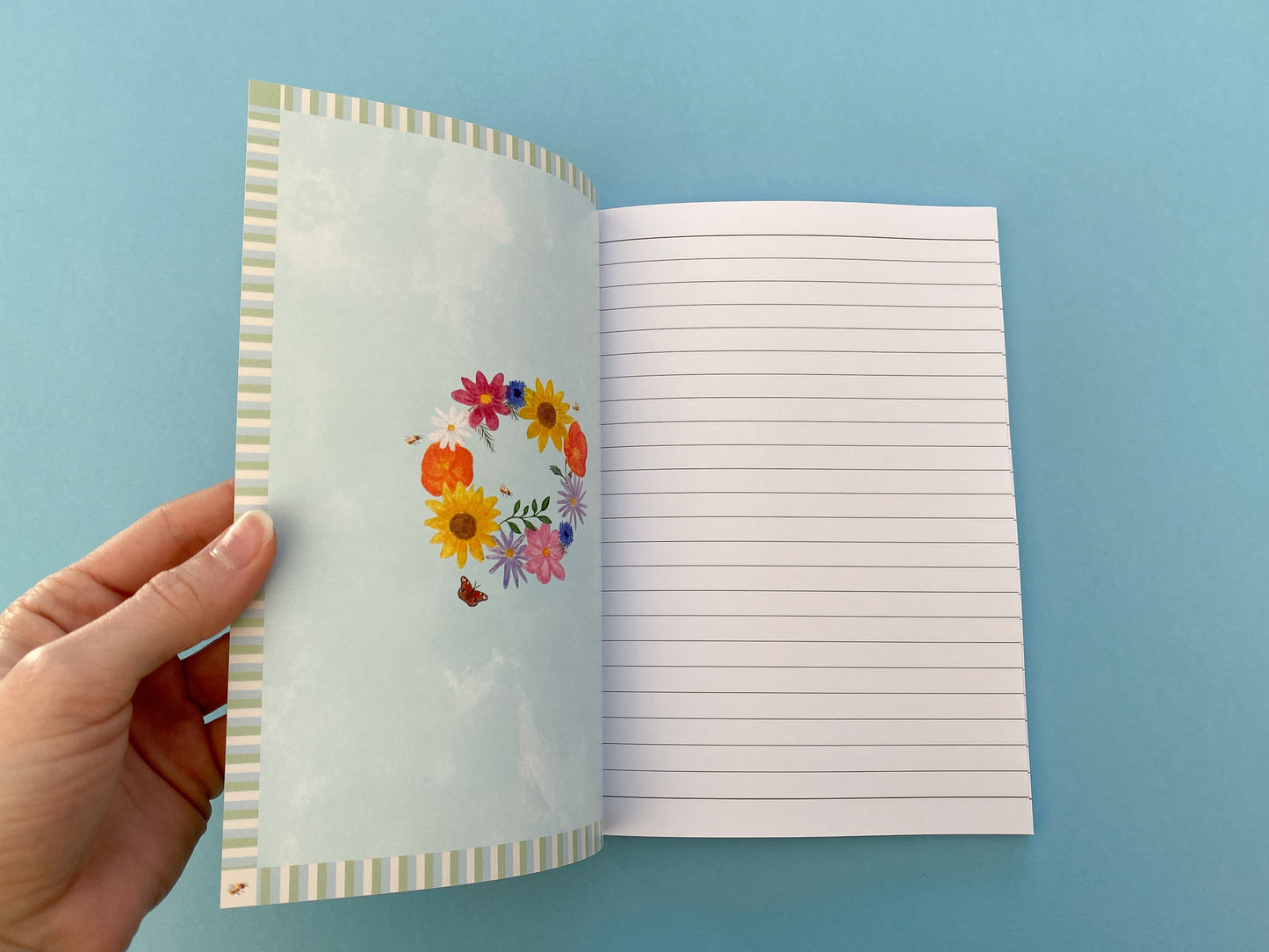 Inside front and back cover design. Stripy pattern border. Pale blue watercolour background with summer wreath in the middle. Flowers include pink cosmos, blue cornflowers, Sunflowers,  orange poppies, purple asters, daisy, green foliage, bees and a peacock butterfly.