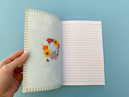 Inside front and back cover design. Stripy pattern border. Pale blue watercolour background with summer wreath in the middle. Flowers include pink cosmos, blue cornflowers, Sunflowers,  orange poppies, purple asters, daisy, green foliage, bees and a peacock butterfly.